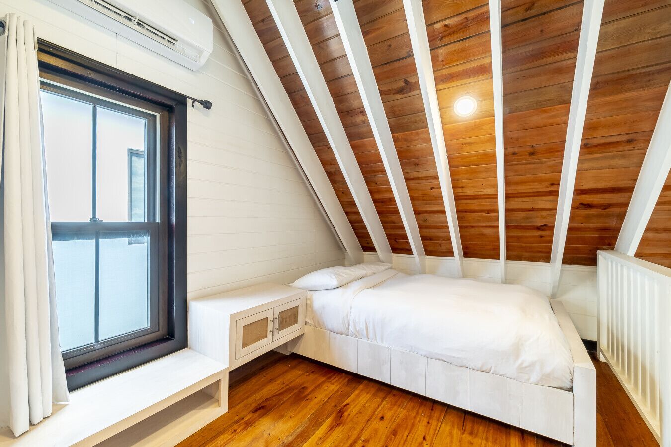 Ein kleines Schlafzimmer mit geneigtem Dach, einem Fenster und einem weißen Bett, inklusive eingebauter Sitzbank.
