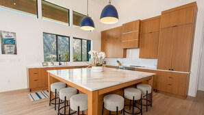 A modern kitchen with wooden cabinets, a marble island with bar stools, pendant lighting, and a view of mountains through large windows. - Private Pool | Stunning lake & mountain views | Modern, costal design (Manson)