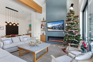 A modern living room with a fireplace, a large Christmas tree, and a television mounted on the wall. - Private Pool | Stunning lake & mountain views | Modern, costal design (Manson)