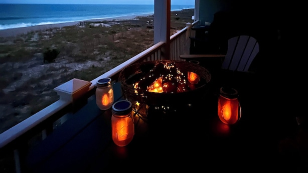 A balcony with a view of the beach, a fire pit, and string lights.