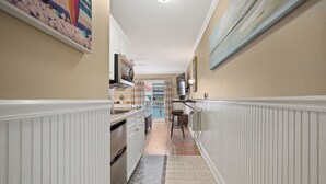 Un pasillo estrecho con paneles de madera blanca, una zona de cocina con microondas y armarios, y un comedor con mesa y sillas.