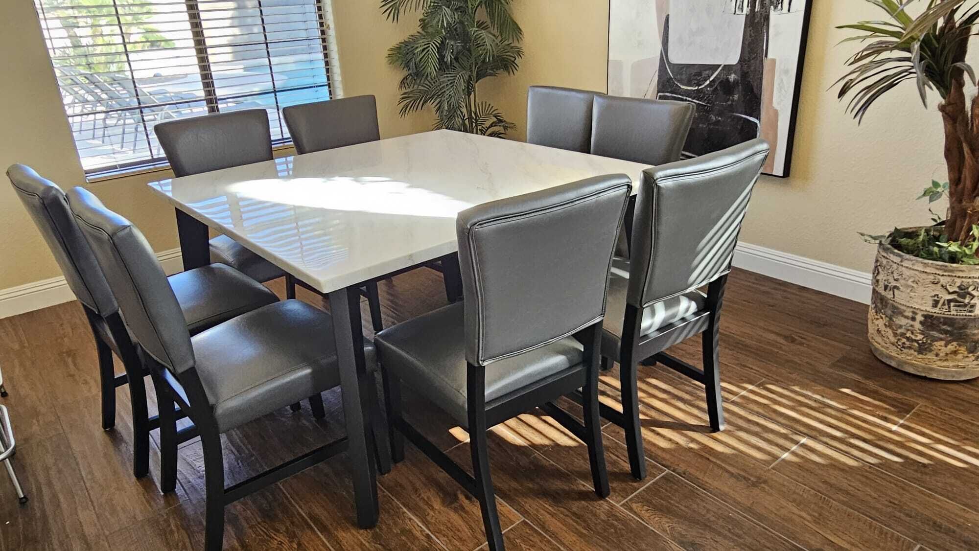 A dining area with a rectangular table surrounded by six chairs, a potted plant, and a window with blinds.