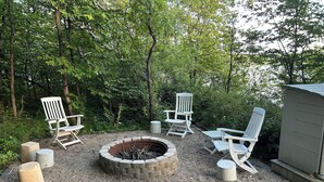 Ein Lagerfeuer, umringt von Adirondack-Stühlen, einer Holzterrasse und einem Schuppen in bewaldeter Landschaft.