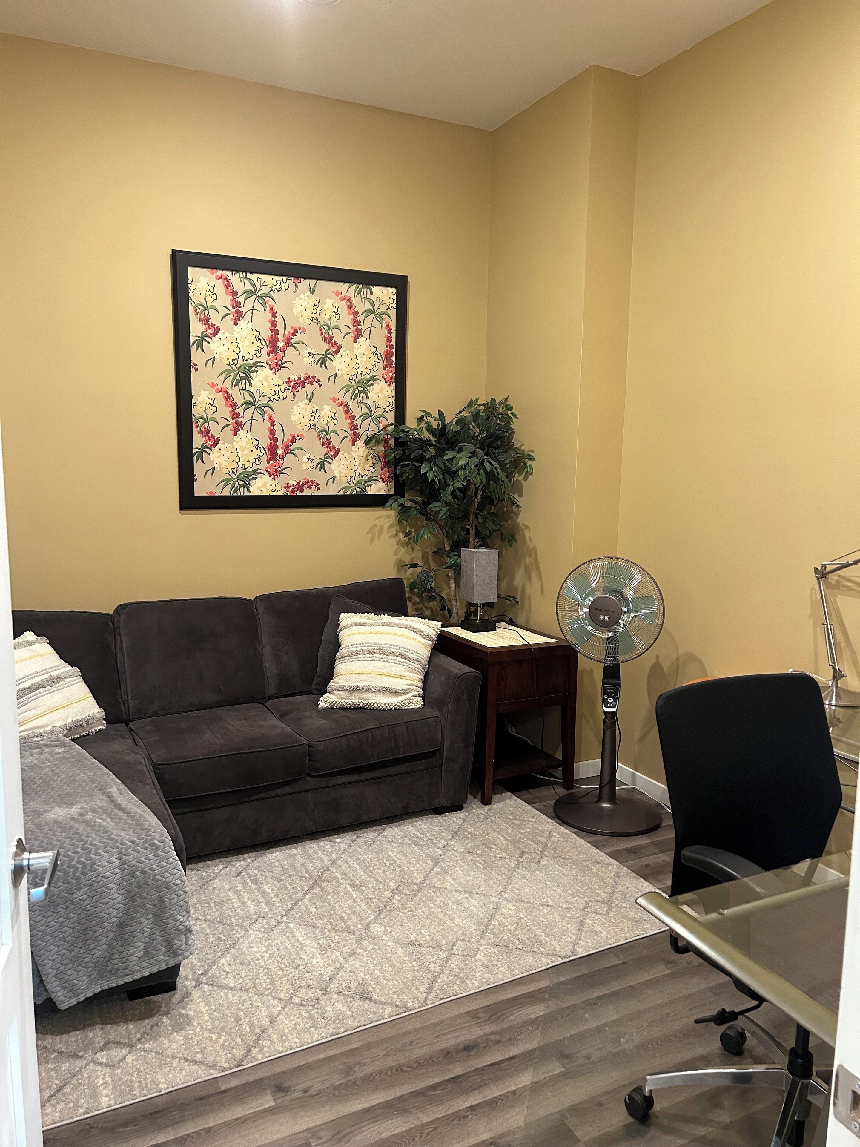 Un salon avec un canapé foncé, une petite table, un ventilateur sur pied, une plante en pot et une œuvre d’art florale encadrée accrochée au mur.