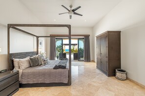 Quarto espaçoso com cama de dossel, ventilador de teto e vista para um pátio com mobiliário de exterior.