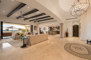 Uma sala de estar ampla, com um sofá grande, uma mesa de centro e vista para o oceano por uma porta de vidro.