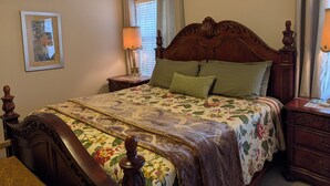 A bedroom with a wooden bed, floral bedding, a nightstand with a lamp, a framed picture on the wall, and a window with curtains.