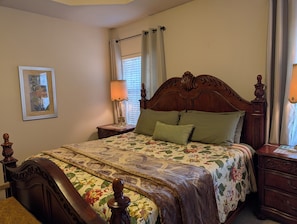 A bedroom with a wooden bed, floral bedding, a nightstand with a lamp, a framed picture on the wall, and a window with curtains.