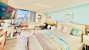 A modern hotel room with a large bed, a desk with a computer, a chair, and a view of the city through a large window. - Waikiki Ocean&Sunrise View Studio (Honolulu)