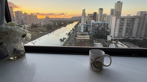 A view of a cityscape with tall buildings and a river, viewed from a window with a mug on the windowsill. - Waikiki Ocean&Sunrise View Studio (Honolulu)