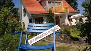 Una silla azul con un cartel que dice “JUST ANOTHER DAY IN PARADISE” frente a una casa con techo rojo y balcón.