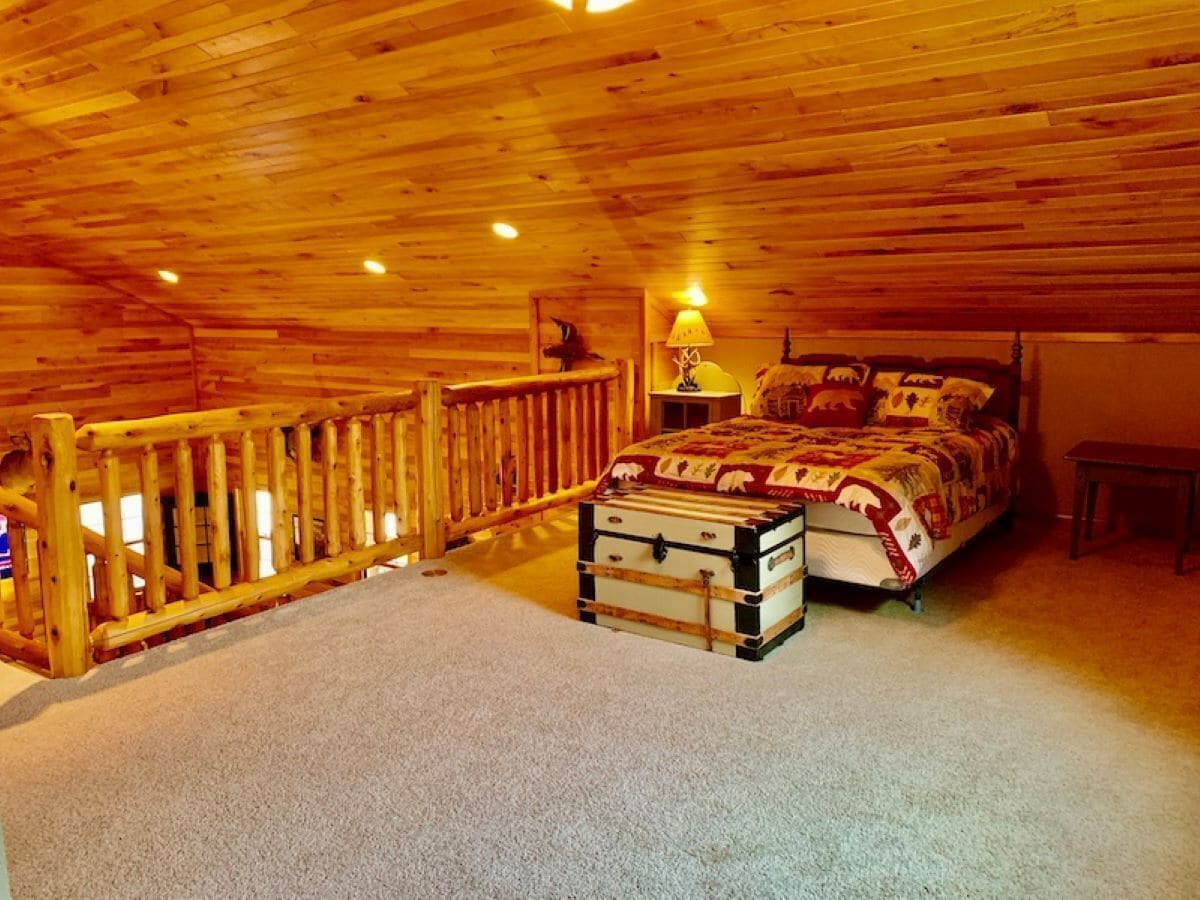 A bedroom with a wooden ceiling, a bed with a patterned blanket, a suitcase, and a nightstand with a lamp.
