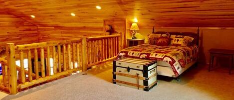 A bedroom with a wooden ceiling, a bed with a patterned blanket, a suitcase, and a nightstand with a lamp.