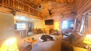 A spacious living area with a high ceiling, wooden beams, a dining area, and a cozy seating arrangement. - Log Cabin/ w hot tub, outdoor pool & pool table (Lansing)