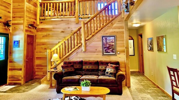 A spacious log cabin interior with a wooden staircase, a cozy living area with a sofa and coffee table, and a ceiling fan. - Log Cabin/ w hot tub, outdoor pool & pool table (Lansing)