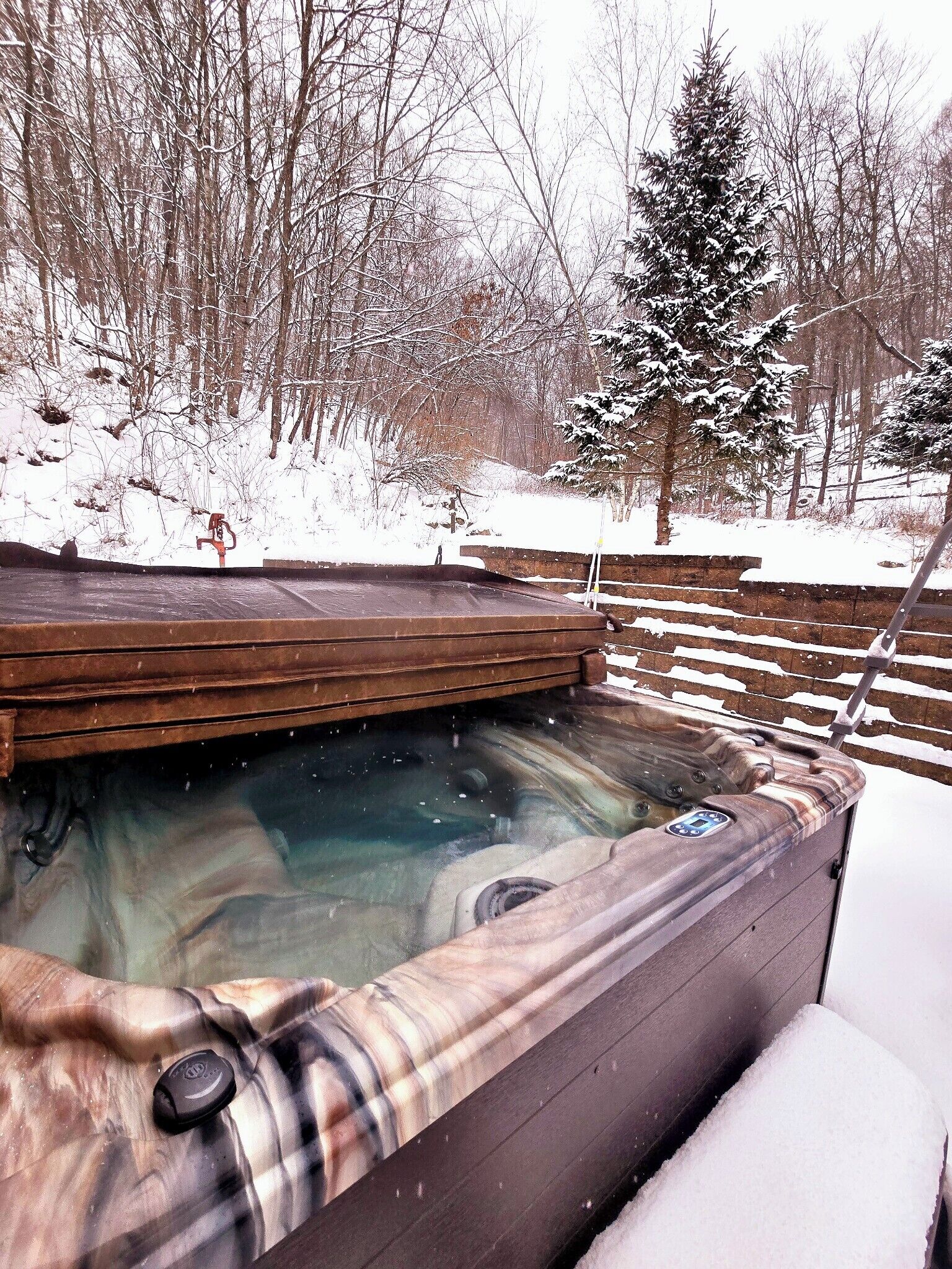 Een hottub met een houten buitenkant, omgeven door een besneeerd landschap en een houten hek.