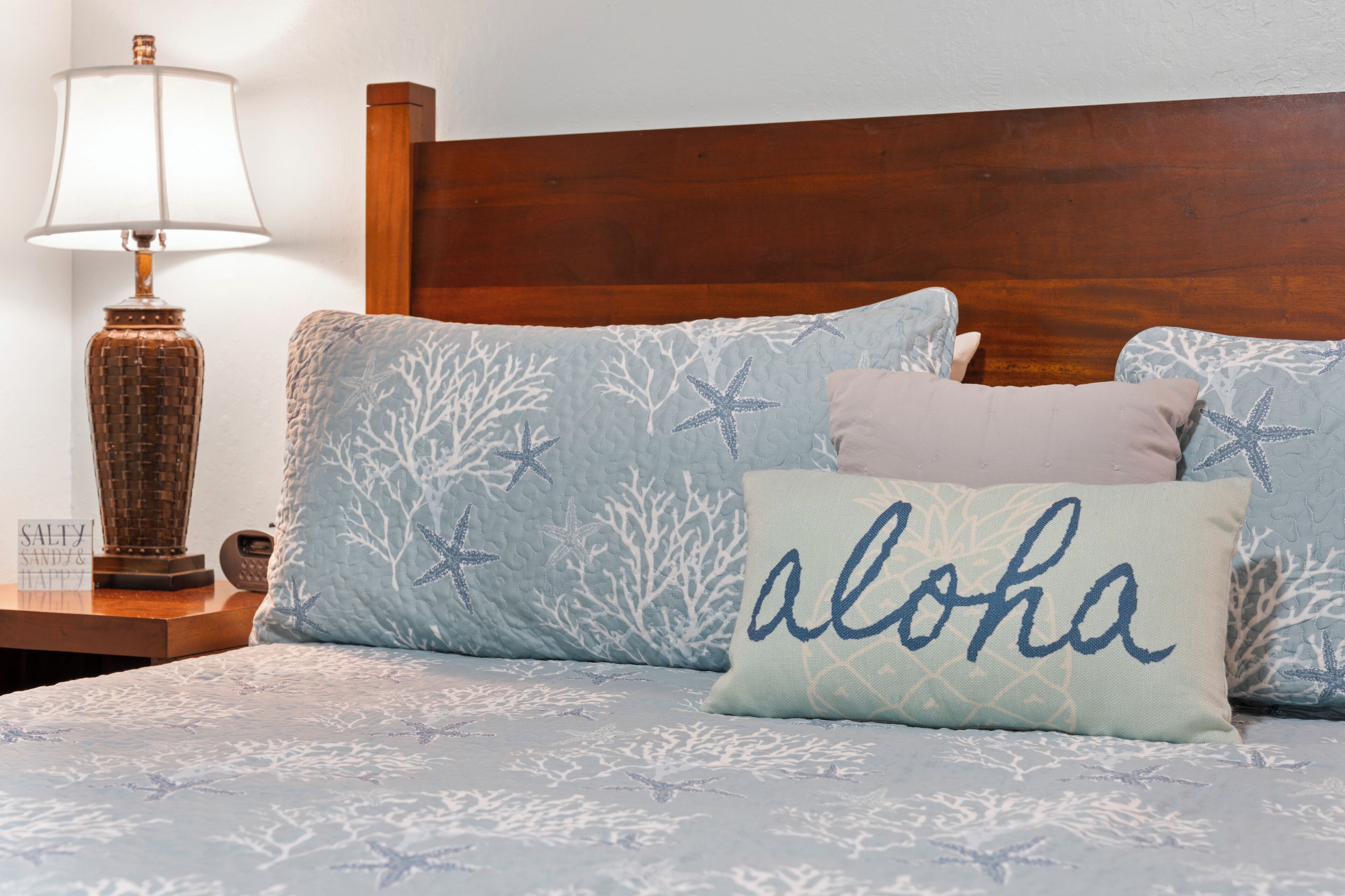 A bed with a blue and white patterned bedspread, a wooden headboard, and pillows with starfish and 'aloha' designs.