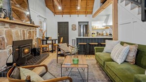 A cozy living room with a stone fireplace, wooden ceiling beams, a green sofa, and a kitchen area in the background. - Corner Cabin • AC • Spa • Game room • Dog Friendly (Big Bear)