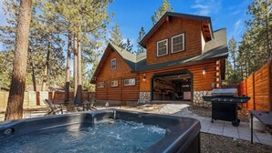 A log cabin with a hot tub, barbecue grill, and outdoor seating area surrounded by trees. - Corner Cabin • AC • Spa • Game room • Dog Friendly (Big Bear)