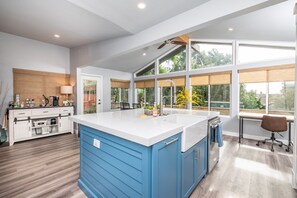 A modern kitchen with a large island, white countertops, and a blue cabinet. There is a dining area with a table and chairs, and a window with a view of greenery.