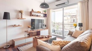 A modern living room with a flat-screen TV, wooden shelves, a sofa, and a large window with a city view.