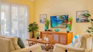 A living room with a wooden coffee table, two white sofas, a TV on the wall, and a large window with curtains. - Beachfront residence in central Sosua, 2BD/2BTH with sea views. Chica-friendly! (Sosúa)