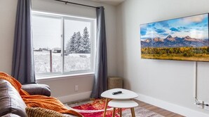 Ein Zimmer mit großem Fenster, grauen Vorhängen, einem grauen Sofa, einem kleinen weißen Tisch und einem Gemälde von einer Berglandschaft.
