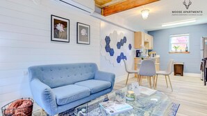 Een moderne woonkamer met een blauwe bank, een glazen salontafel en een keuken op de achtergrond.