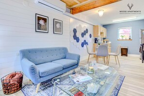 A modern living room with a blue sofa, a glass coffee table, and a kitchen area in the background.