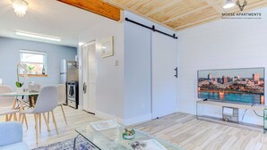 Een moderne woonkamer met een glazen salontafel, een eethoek met stoelen, een keuken met inbouwapparatuur, en een grote televisie waarop een stadsgezicht te zien is.