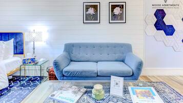 A modern living room with a blue sofa, a glass coffee table with books, a lamp, and framed pictures on the wall.