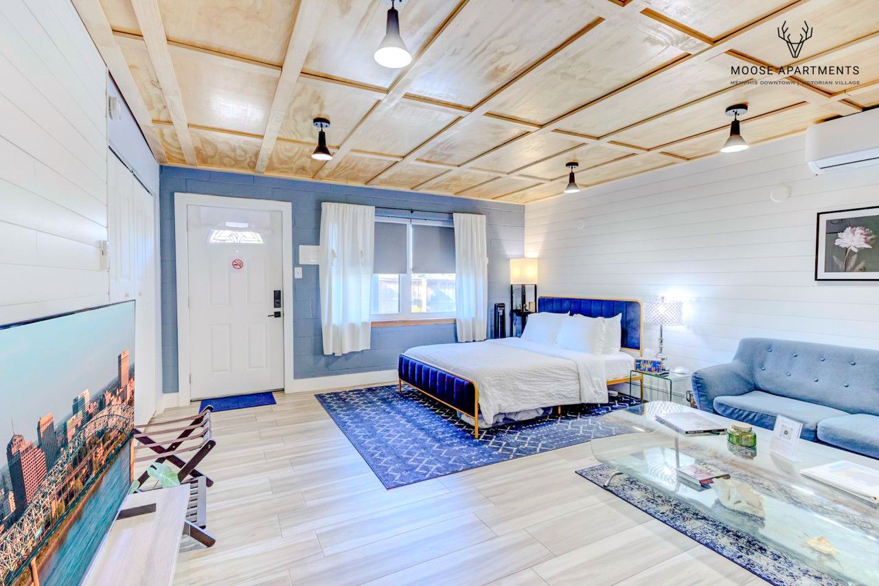 Un dormitorio moderno con cama, un sofá azul, una mesa de centro de vidrio y una televisión con vistas a la ciudad.