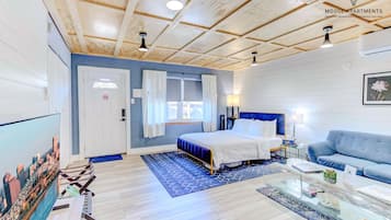 Un dormitorio moderno con cama, un sofá azul, una mesa de centro de vidrio y una televisión con vistas a la ciudad.