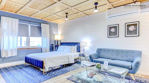 Een slaapkamer met een bed, een blauwe slaapbank, een glazen salontafel en een raam met gordijnen.