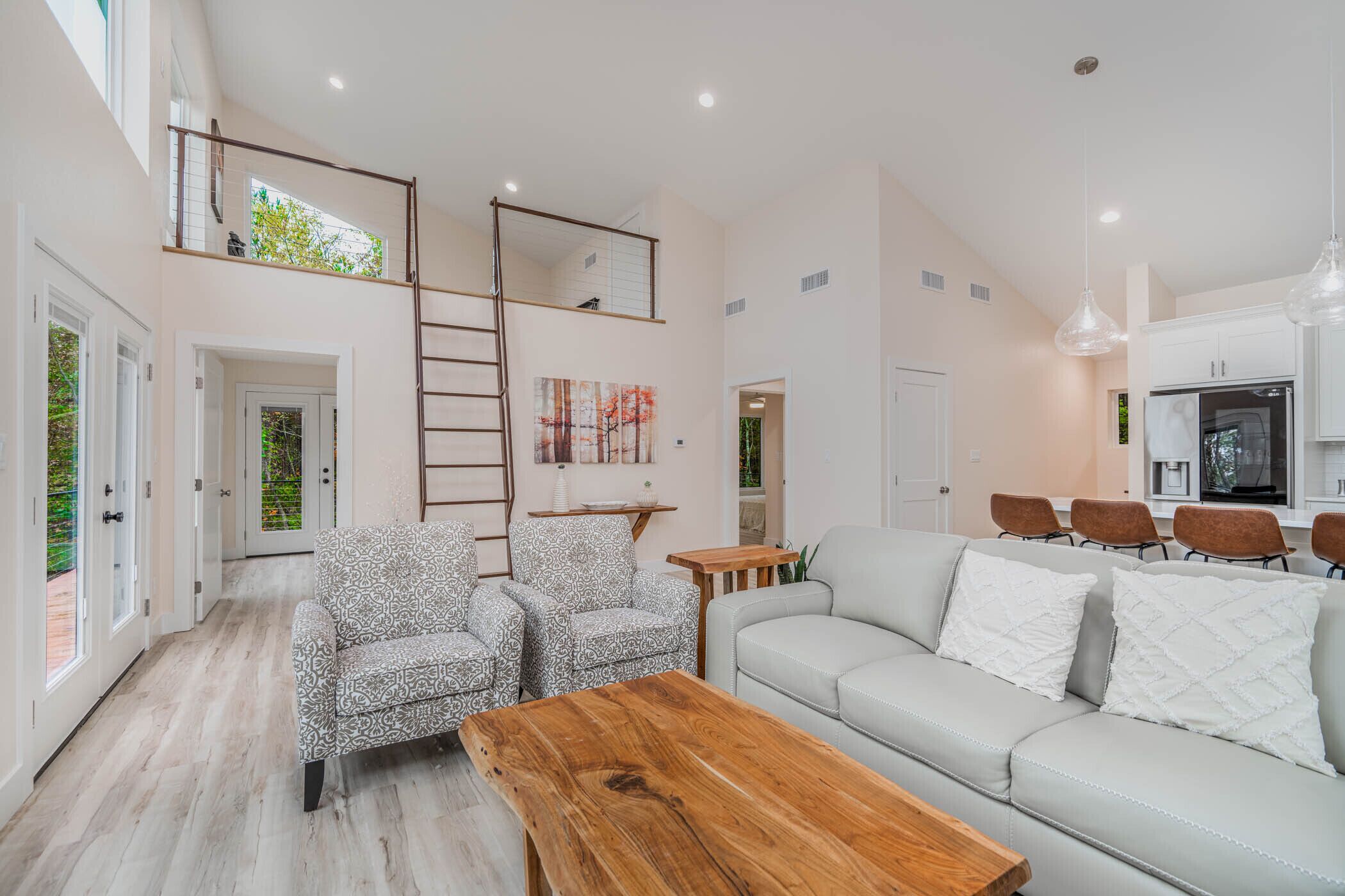 Un salón espacioso con techo alto, una mesa de centro de madera, sillones estampados, un sofá blanco y una zona de cocina con electrodomésticos modernos.
