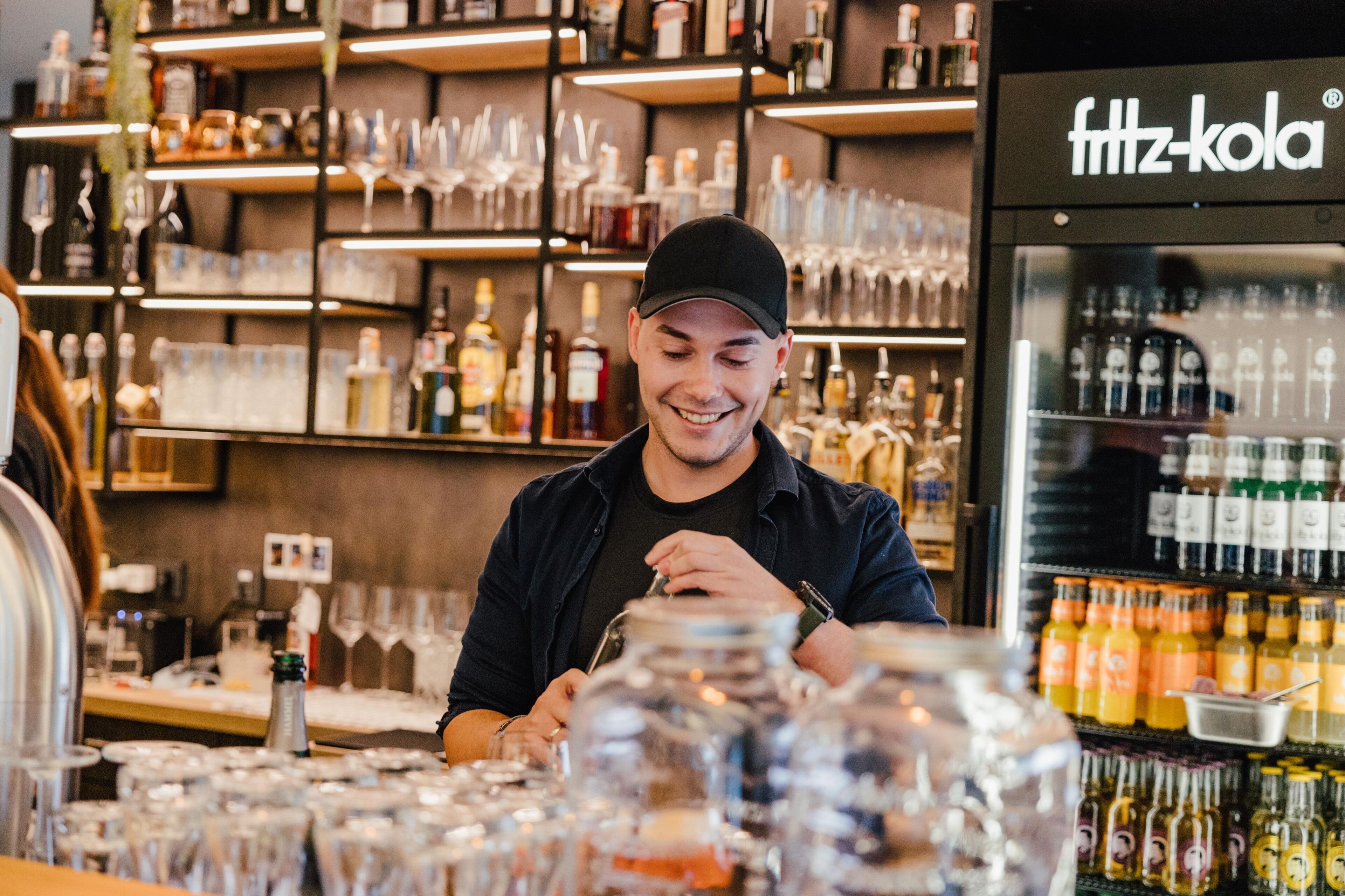 Una zona de bar con estantes llenos de botellas y vasos, y una vitrina refrigerada con botellas.