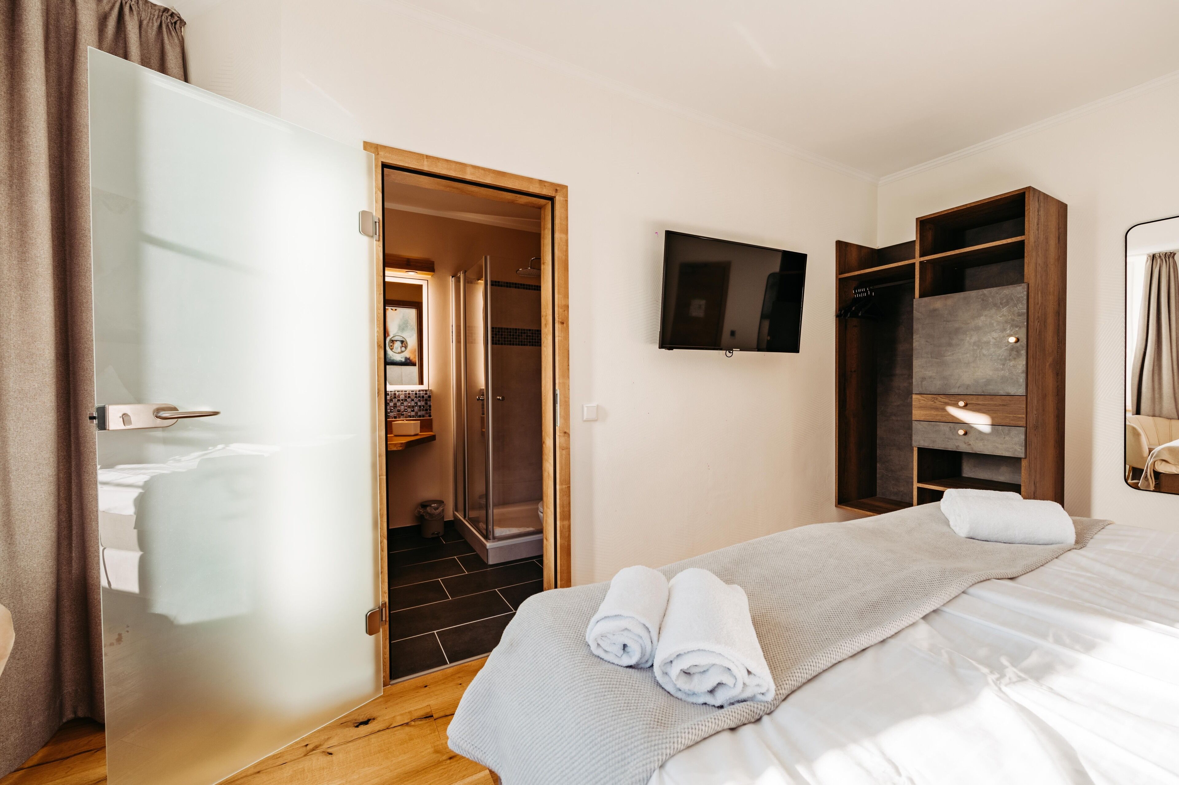 A hotel room with a bed, a TV mounted on the wall, a wooden wardrobe, and a shower area.