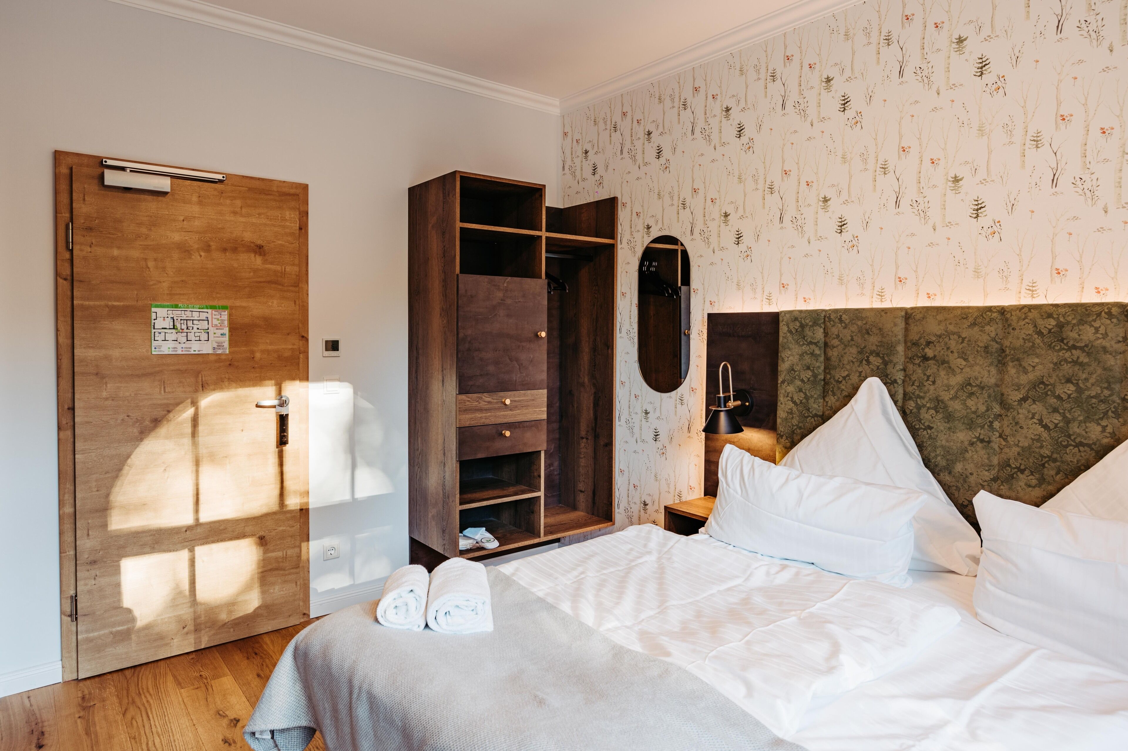 A hotel room with a wooden wardrobe, a bed with white linens, and a floral wallpaper pattern.