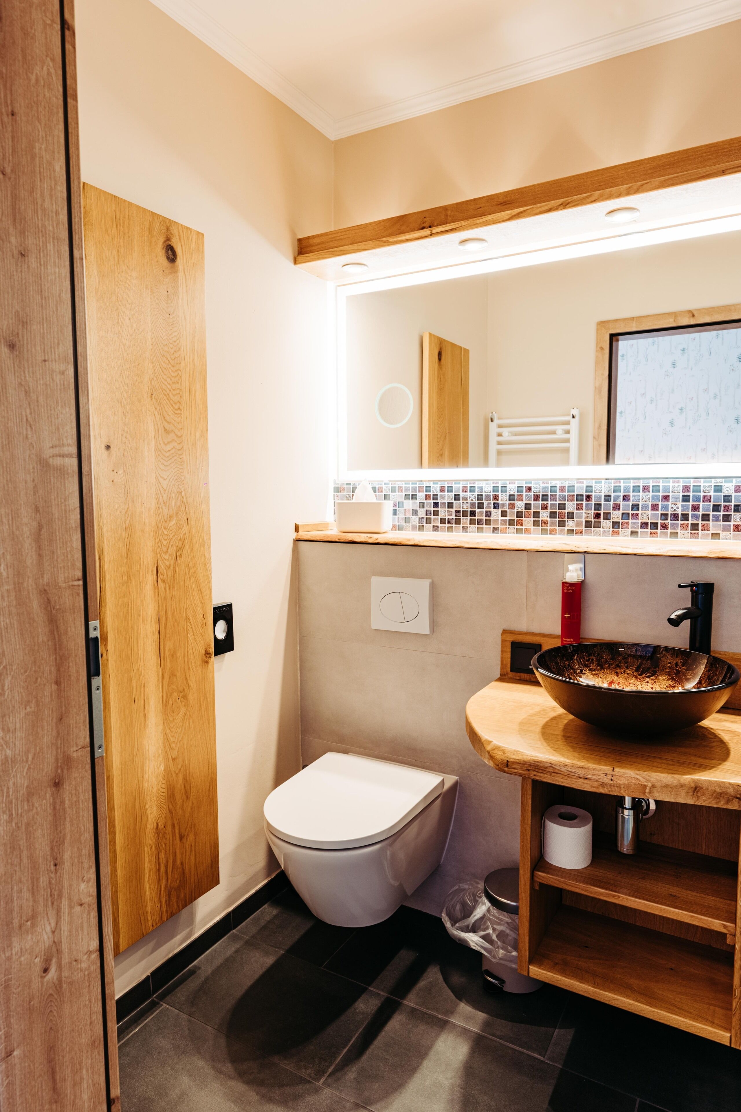 Een badkamer met een houten wastafelmeubel, een grote spiegel, een wit toilet en een betegelde spatwand.