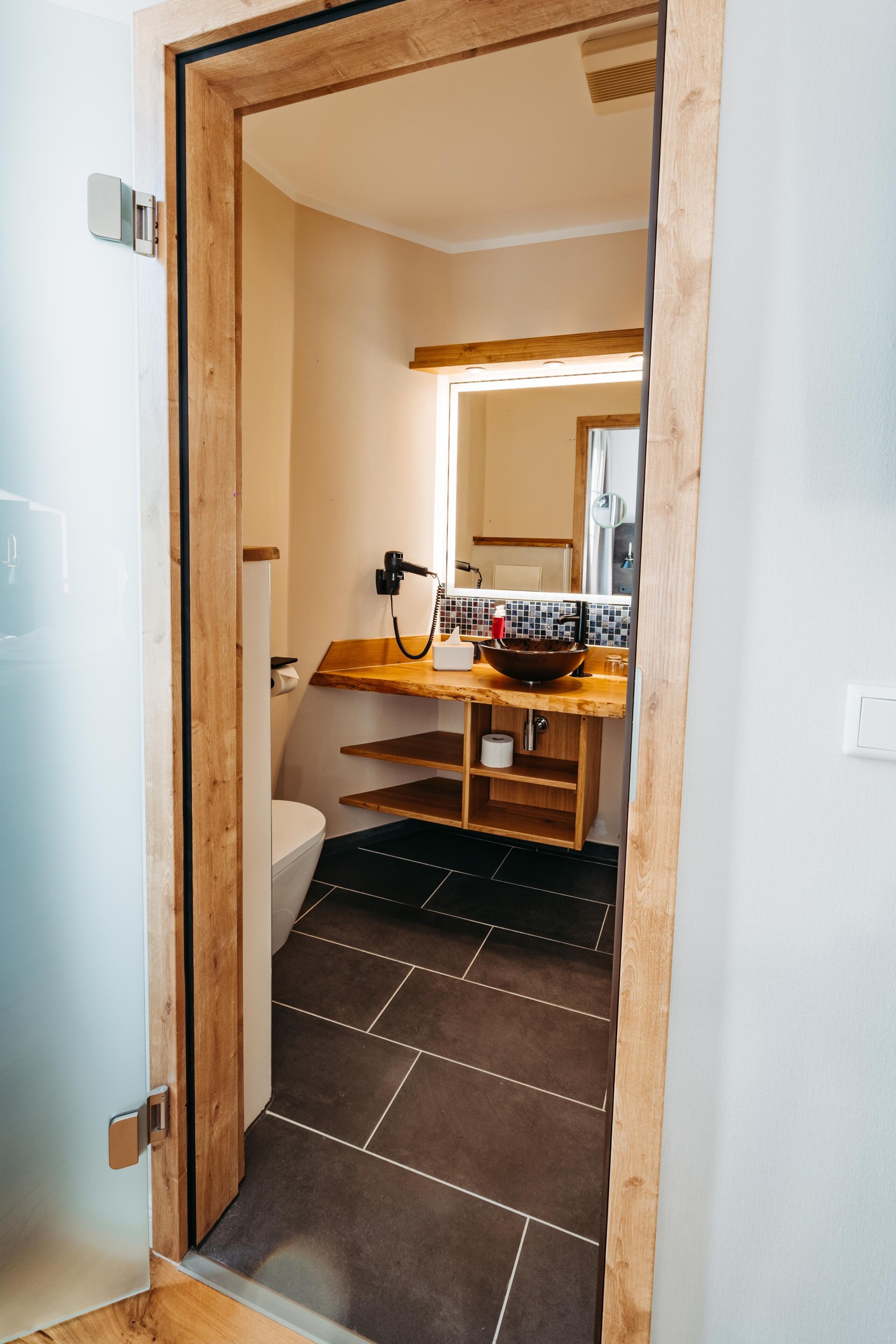 Ein Badezimmer mit Holztür, Toilette, Waschbecken mit Spiegel und Fliesenboden.