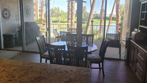Un comedor con una mesa y sillas, un reloj en la pared, y vistas a un campo de golf y palmeras a través de amplios ventanales.