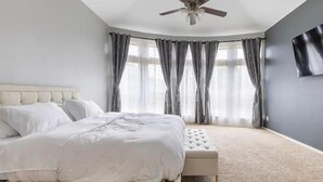 A bedroom with a bed, a ceiling fan, a large window with curtains, and a wall-mounted TV. - Luxury 1 story 3,272 SF. 12 miles from IAH airport (Houston)