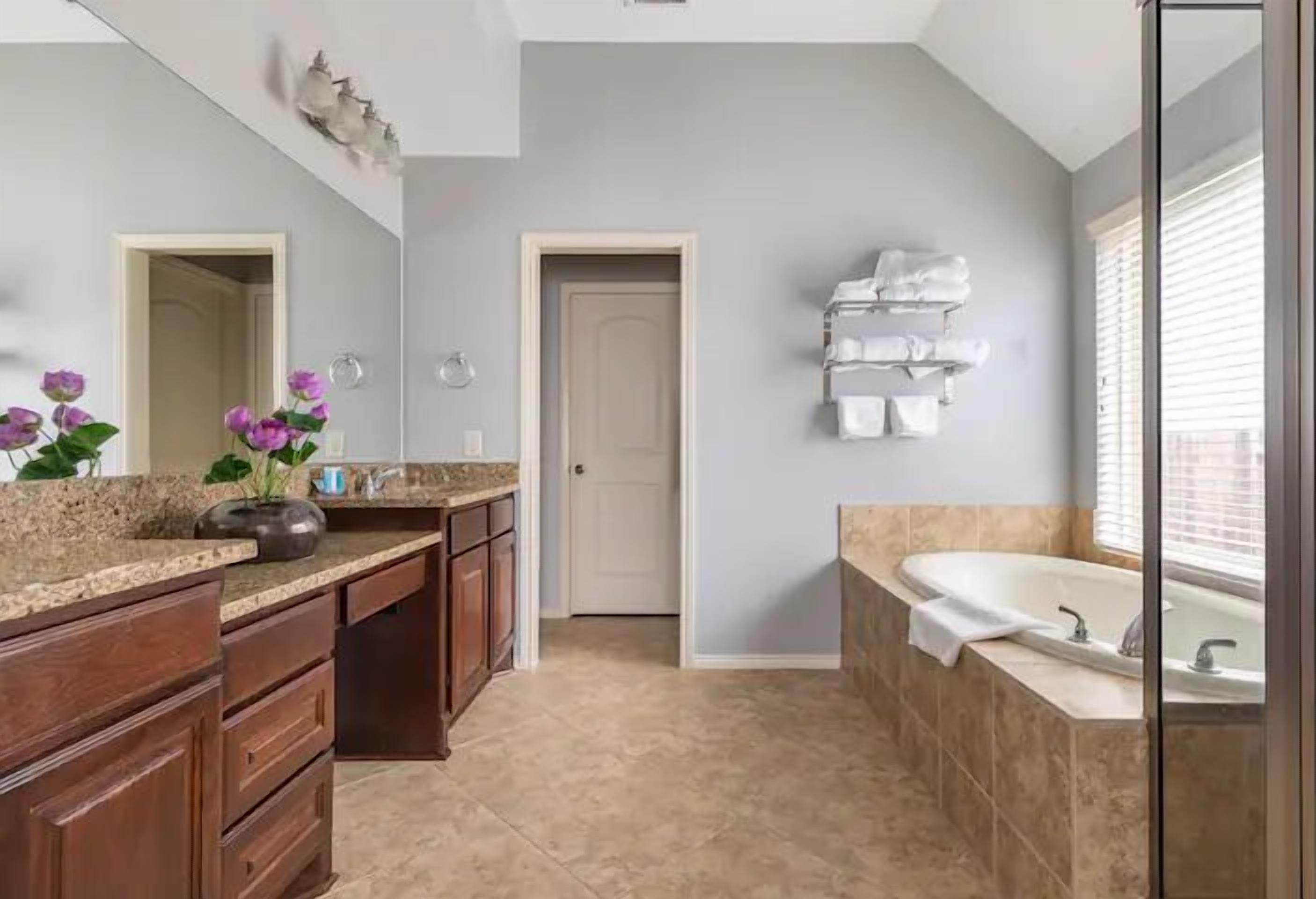 A bathroom with a large bathtub, a sink, and a mirror. There is a towel rack on the wall and a door leading to another room.