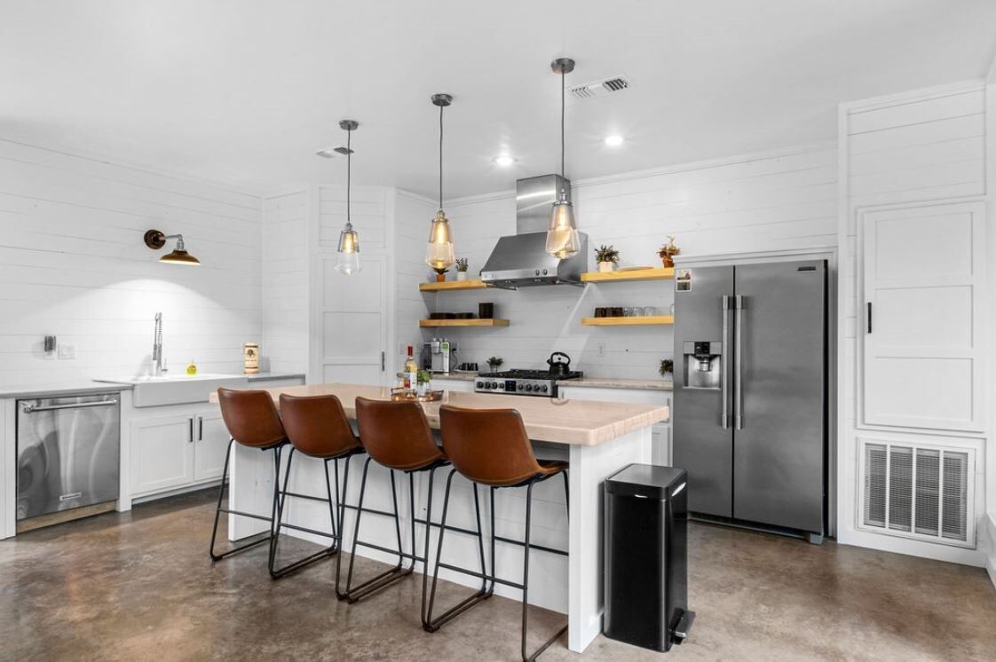 Une cuisine moderne avec des meubles blancs, un îlot central doté de tabourets de bar, des appareils en acier inoxydable et un éclairage suspendu.