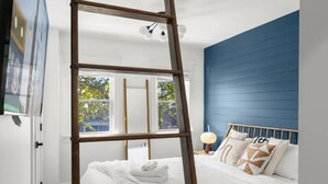 Ein Schlafzimmer mit einer Holzleiter, einem Bett mit weißer Bettwäsche, einer blauen Akzentwand und einem Fenster mit Blick auf Grünflächen.