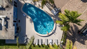 Vista aérea de uma piscina cercada por espreguiçadeiras e palmeiras, com um calçadão pavimentado e um carro estacionado.