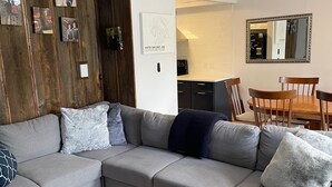 Een moderne woonkamer met een grijze hoekbank, houten wandpanelen aan een muur en een eethoek met een houten tafel en stoelen.