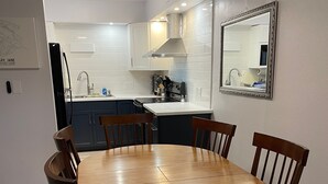 Een eetgedeelte met een ronde houten tafel en vier stoelen, een keuken achtergrond met witte kastjes en een roestvrijstalen afzuigkap, en een spiegel aan de muur.