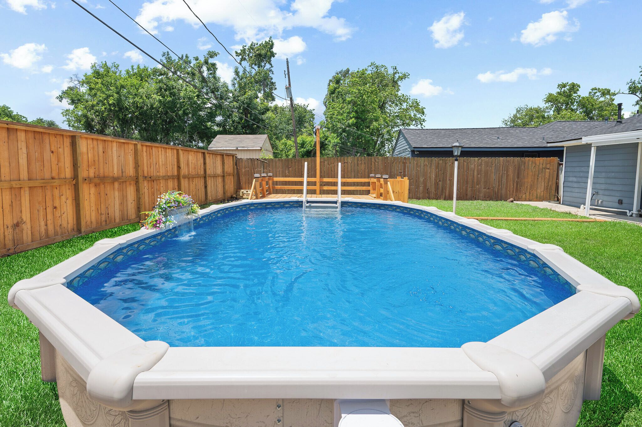 An above-ground swimming pool with a wooden deck and a ladder, surrounded by a wooden fence and a grassy yard.