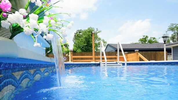 Una piscina con fuente, cerca una valla de madera y una casa al fondo.